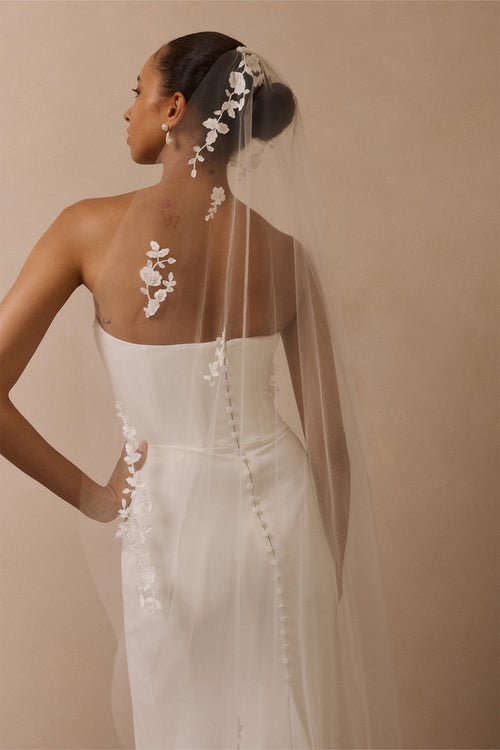 A woman shown from the back wearing a strapless white wedding gown with a sheer tulle veil adorned with white floral applique, against a warm beige studio background.