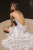 A woman seated on a circular beige platform, photographed from behind, wearing a white lace bridal gown with thin straps, a deep open back, and delicate chain detailing across the upper back.