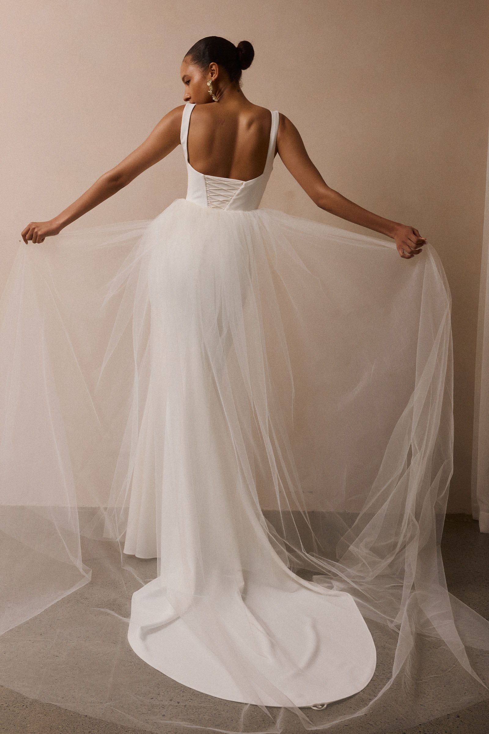 A woman wearing a white wedding dress is photographed from behind, holding out the sheer tulle train against a plain beige studio wall_XS_