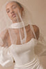 A woman wearing a white bridal gown with a high neck, dramatic puffed sleeves, and a sheer veil, posed against a warm beige background.