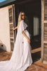 A woman wearing a flowing white dress and sunglasses stands in a doorway with wooden shutters, with warm sunlight casting shadows on the wall and floor.