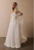 A woman wearing a white lace bridal gown with a long sheer veil and fringe detail, standing in a studio with a warm beige wall and dark floor_XS_