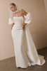 A woman wearing a white off-the-shoulder wedding gown with dramatic puffed sleeves, a fitted bodice, and a long train, styled with a sheer veil against a warm beige wall.