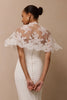 A woman shown from the back wearing a white wedding dress with a sheer lace cape-style back overlay and a row of small back buttons, against a plain beige studio background.
