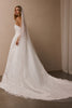 A woman wearing a strapless white wedding gown with a long sheer veil, photographed indoors against a warm beige wall and floor.
