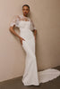 A woman modeling a white wedding gown with a lace high-neck bodice and sheer lace cape sleeves, standing against a plain beige wall with a floor visible.