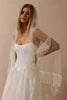 A woman wearing a white lace bridal gown and a long sheer white veil with fringe trim, posed against a warm beige studio background.