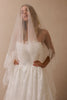 A woman wearing a strapless white lace wedding dress with a long sheer white veil draped over her face and shoulders, posed against a warm beige studio background.