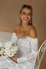 Woman in a lace wedding dress holding flowers against a neutral background
