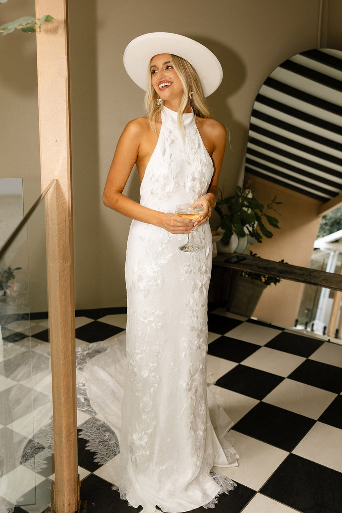 Woman in a halter neck lace wedding dress and hat holding a glass of champagne