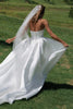 Woman in a white wedding dress with a long train and veil, standing on grass.